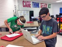 A young teenage boy and girl, in middle school or ninth grade, are working at a classroom table. The boy is working on a HP laptop, and the girl is putting super glue on a piece of paper that's on top of a red lunch tray.