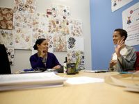 Two female teachers are sitting at a large table in a classroom. They're smiling and talking.