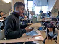 A high school boy is sitting at his desk, smiling, folding a piece of cardboard with pieces of tape on it. The class is filled with students at their desks or on the floor cutting and taping together pieces of cardboard.
