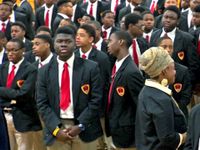 An assembly of teenage boys in tan slacks, white-collared, button-up shirts, black jackets, and red ties are standing. A female teacher is standing among them.