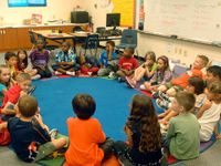 A group of young elementary students are sitting in an oval on their classroom floor on a blue circular rug with a colorful border.