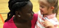 A fifth-grade girl is kneeling on a basketball court, holding a young pre-school girl with special needs. They're both looking at each other and smiling. 