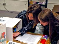 Two young women are squatting by a table in a classroom; one of them is reaching into a cardboard box that's part of a display.