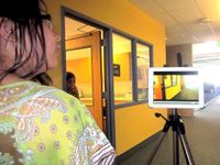 A tripod is standing in the middle of a school hallway with an iPad on it. A student is standing behind it, looking off to the side. Another student is standing between a doorway, looking into the hallway. 