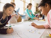 A group of six young students are standing alongside a long table with art paper stretched across it. Each student is wearing a long, white apron and is drawing on the paper with markers.