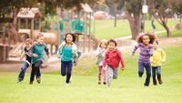 Eight young kids are running outside on the grass in front of a playground.
