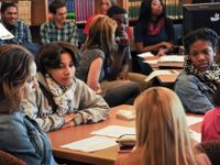 A group of ten high school students are in a library sitting at tables and talking to each other.