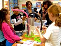 Three elementary-aged girls and two female teachers are standing around three rectangular desks pushed together with a student-made contraption standing on the center desk. The contraption is a large, circular, aluminum-looking tube standing about two to 