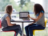 Two teenage girls are sitting at a small circular table with a textbook laid out, looking at a laptop. They're sitting next to a glass wall that looks out to the school parking lot.