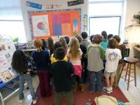 A group of elementary students are huddling together in class, looking at a pie chart and bulletin board on the wall.