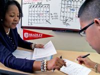 An adult female in a counseling room is sitting across a small table from a teenage student, helping him fill out a form.