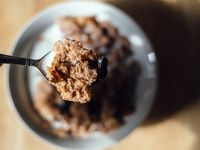 A closeup of a spoon with milk, corn flakes, and raisins. The rest of the image is out of focus. Beneath the spoon is a bowl filled with cereal on a light brown, wooden table. 