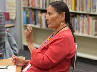 A side profile closeup of a female teacher sitting at a table in a classroom. A bookshelf behind her, and she's talking to someone. She's wearing a red shirt, a long, beaded necklace, and circular earrings.