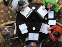 An aerial shot of five teachers sitting around a dark brown circular table in light brown cloth-covered chairs. There are three laptops, four notebooks, and three cups of coffee on the table.