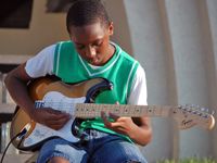 A young boy in jean shorts, a white t-shirt, and a green sweater vest is sitting outside in front of his school playing an electric guitar.