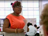 A teenage girl is standing in a classroom, holding a piece of paper, speaking to a small group of people, while other students are grouped throughout the classroom.