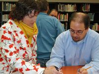 A woman in a floral sweater and a yellow scarf is standing, pointing to a piece of a paper on a table with her pen, and a man wearing glasses and a blue button-up shirt is sitting beside her, looking at the same paper. A bookshelf is behind them.