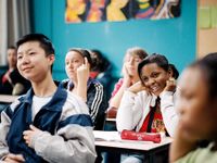A group of high school students are sitting at desks in class, all looking towards the front, smiling.