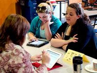 Three female teachers are sitting at a table, two on one side and one on the other, talking to each other and looking at their tablets.