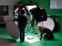 Three students and a teacher are in a green room, setting up the lighting to film.