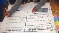 A student is drawing on a big piece of paper, filling in two columns, "Supporting Details" and "Evidence From."