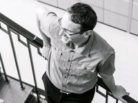 A male teacher in glasses is leaning against a stairway rail, smiling. 