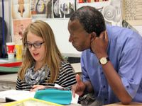 A male teacher is sitting next to a female student. The student is pointing to a page on an open textbook, talking. There is artwork on the wall behind them.