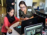 A female adult wearing a green shirt and glasses is sitting beside a young female student in a red blouse. They're both looking at a laptop computer.