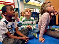 A group of young students are sitting on the floor in their classroom, looking up.