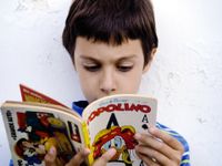 A closeup of a young boy in a blue striped shirt against a white wall reading a book