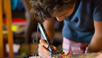 A closeup of a teenage boy bent over, holding a long electronic device, connecting wires to metal pegs on a wooden board.