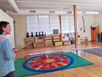 A female teacher is standing at the left side of a classroom. The classroom is empty, except for a large green and brown rug placed side by side with two benches placed on top of them, and shelves are against the back wall with drums on top of them.