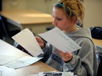 A teenage girl in a gray sweatshirt is sitting at her paper-scattered desk, holding pieces of paper in both hands.