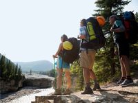 Three hikers carrying backpacks with helmets and bed rolls attached, standing looking down at a creek