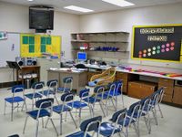 17 chairs are lined side-by-side in three rows facing the front of a classroom. At the front of the class, inches from the ceiling, a flat screen TV is attached to the wall. A countertop with drawers extends along the right side of the room.