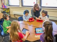 Five young elementary students are sitting around a six-sided table in a classroom. They're all wearing headphones and playing on iPads. A female teacher is assisting one of them.