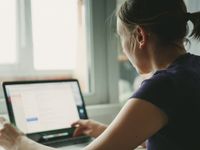 A closeup of a young woman, facing away from us, sitting down next to a window, typing on her laptop. Light is shining into the room from the window.