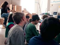 A group of 8 students are sitting cross-legged on the classroom floor. One student is sitting on what looks like a student-made chair out of squares of wood. Buildable blocks of wood occupy one side of the classroom.
