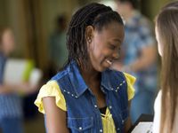 A teenage girl in a yellow shirt and jean vest is smiling, looking down at an opened binder in her hands, showing it to another girl outside of the frame of the photo. The rest of the photo is blurred out, and seems to be students in a school hallway.