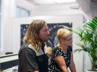 A side profile of a male and female adult from the waist up standing in a spacious white-walled room looking in the same direction. There's a black and white painting behind them, and a green, potted plant beside them.