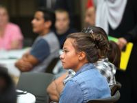 A teenage girl is sitting at a table among other students sitting at tables in a classroom. Everyone else is blurred out.