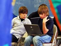 Two young teenage boys are sitting in foldable chairs in class. One has a laptop on his lap that they're both looking at.