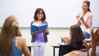 A young girl is smiling, standing at the front of the class and reading, and her teacher and peers are clapping for her.