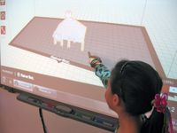 A closeup of a young girl standing next to and pointing at a projector screen. A 3D drawing made up of small squares is projected on the screen, showing the inside of a white room with a brown floor and a small white pyramid in the middle.