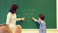 Elementary school teacher works with student on math problem on chalk board in classroom