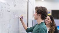 High school student writes math equation on board with teacher