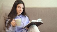 Teen girl reads a book while sitting on a couch and drinking from a mug
