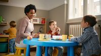 Preschool teacher sits with students eating a snack