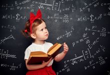 A toddler in a red and white outfit holding an open book in front of a blackboard covered in advanced math formulas