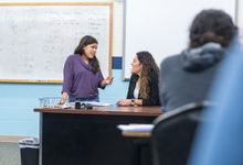 High school student speaking with teacher at teacher;s desk in classroom. 